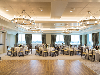 A bright reception room arranged with round tables, white linens, soft floral centerpieces, and expansive windows overlooking the surrounding area.